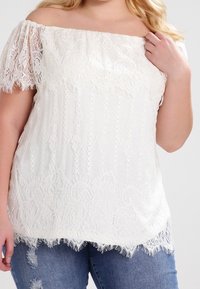 Haut en dentelle blanche à épaules dénudées avec des manches courtes, présentant des motifs complexes et un ourlet festonné. Associé à un jean en denim déchiré.