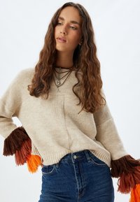 Maglione beige lavorato a maglia con maniche lunghe e frangette nei toni di marrone e arancio, dotato di un ampio scollo e una vestibilità rilassata. Abbinato a jeans blu.