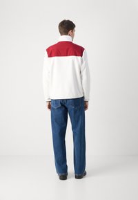 Giacca bianca con un pannello rosso sulla spalla, colletto alto e polsini a costine, abbinata a jeans blu e scarpe nere. Vista posteriore.