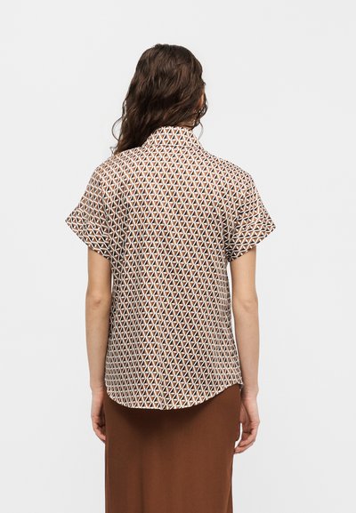 Γυναίκα με μακριά σκούρα μαλλιά που φορά κοντομάνικο πουκάμισο με γιακά και καφέ-λευκό γεωμετρικό σχέδιο και καφέ φούστα, φαίνεται από πίσω.