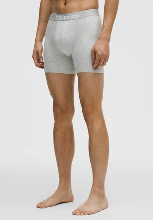 Modello maschile che indossa boxer grigio chiaro Lululemon, in piedi a piedi nudi su uno sfondo bianco uniforme.