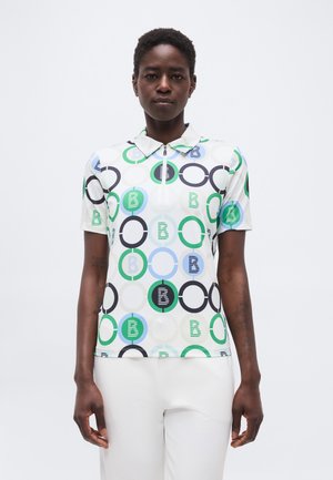 Moteris dėvi trumpomis rankovėmis polo marškinėlį su žaliais, mėlynais, juodais ir baltais apskritimais bei baltas kelnes, stovinti priešais paprastą foną.