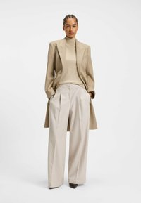 Beige oversized blazer, ribbet turtleneck og brede lyse bukser. Glat stof, skræddersyet pasform, subtile plisseringer og spidse sorte hæle.