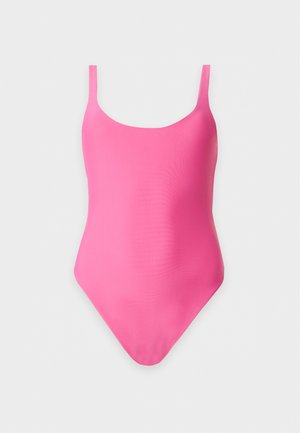 Vrouw met nat haar in een roze badpak, staand op blote voeten in profiel tegen een effen lichte achtergrond.