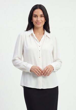 Kvinde med langt mørkt hår iført hvid bluse, lagdelte guldkæder og sort nederdel, stående mod en enkel lys baggrund, hænderne foldet.