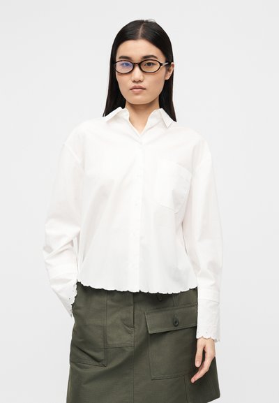 Λευκό πουκάμισο με κουμπιά, με κομμένο μήκος και δαντελένιο τελείωμα με κυματιστές άκρες. Συνδυάζεται με πράσινη φούστα χρηστικότητας με μπροστινές τσέπες.