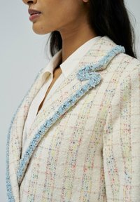 Blazer de tweed blanco con motas multicolores, con un suave ribete texturizado azul claro en el cuello y el frente, complementando una camisa blanca debajo.