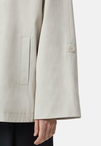 Veste imperméable beige avec une manche droite et large, présentant un bouton au poignet et une petite poche latérale, fabriquée à partir d'un tissu lisse et texturé.