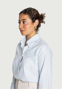 Camicia a righe blu chiaro con colletto, abbinata a pantaloni beige a vita alta. Orecchini dorati e capelli texturizzati completano il look.