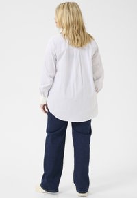 Camisa blanca de manga larga y botones con dobladillo curvado, que presenta un yugo trasero y sutiles rayas verticales, combinada con pantalones de mezclilla de pierna ancha en azul oscuro.