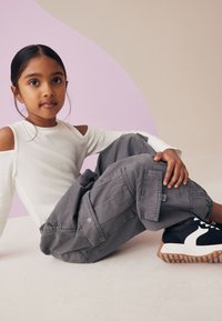 Grå cargo bukser med flere lommer, parret med en hvid langærmet top med udskæringer og sorte sneakers med hvide detaljer, på en lys baggrund.