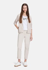 Un cardigan zippé beige clair sur un T-shirt blanc avec des lettres noires et vertes, associé à un pantalon kaki clair et des baskets blanches.