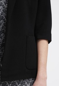 Černý blazer s krátkými rukávy, hladká textura, s přední kapsou. Pod ním je viditelný bílý a černý paisley vzor.