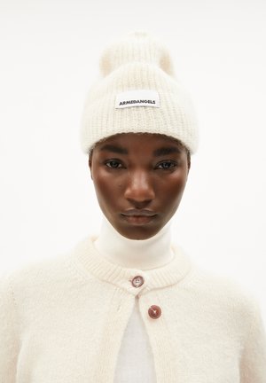 Bonnet en tricot crème avec une texture douce et un pompon rond, présentant une étiquette noire avec le texte "ARMEDANGELS".