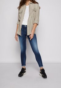 Camisa de botones gris claro sobre una camiseta blanca, combinada con jeans ajustados azul oscuro y zapatillas negras con detalles dorados.