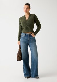 Pulover tricotat de culoare verde măsliniu, scurt, cu margini ondulate, asortat cu blugi din denim bleumarin, cu picioare late, și o geantă maro cu accente aurii.