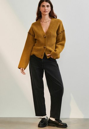 Mustard knit cardigan with a V-neck, button closure, and ribbed texture. Paired with high-waisted black denim and black shoes.