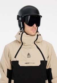 Beige en zwarte sneeuwjas met een hoge kraag, ritsdetails en een logo. Ggedragen met een zwarte helm en grote gemirroredgoggles, allemaal op een witte achtergrond.
