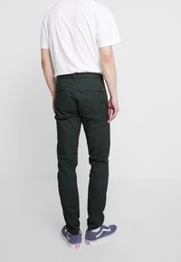 Tmavě zelené, na míru šité kalhoty s pasem a dvěma zadními kapsami na knoflíky, doplněné bílým tričkem a modrými teniskami.