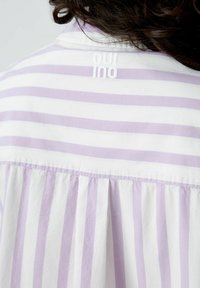 Striped shirt featuring white and light purple colors, with pleated back detail and a collar. Logo embroidered at the neck in white.