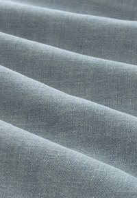 Tissu en denim bleu clair avec une texture lisse, présentant de subtils motifs de tissage diagonal et des plis horizontaux distincts.