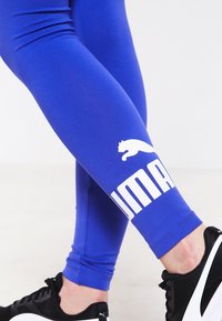 Blå tights i elastiskt material, med en vit Puma-logotyp och varumärkesnamn på underbenet. Kopplade med svarta atletiska skor.