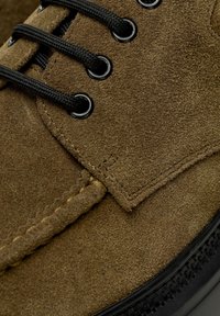 Chaussure en daim marron avec des lacets ronds noirs passés dans des œillets en métal et des coutures détaillées sur le dessus.