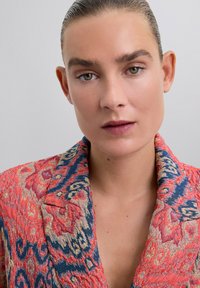 Blazer colorido con una mezcla de patrones rojos y azules, con cuello chal y tejido texturizado. Los detalles incluyen diseños intrincados.