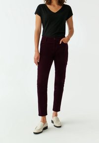 Pantalons en velours côtelé bordeaux à coupe ajustée, poches latérales et fermeture à bouton, associés à un haut noir à col en V et des chaussures beige à enfiler.