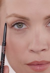 Crayon à sourcils noir tenu près du sourcil, avec les étiquettes "BROW WIZ" et "STRAWBURN" visibles. Peau lisse, couleur de lèvres neutre et yeux clairs.
