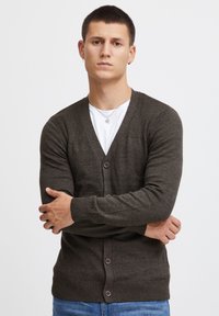 Mørk grønn cardigan med V-hals, ribbet skulderdetaljering, knappelukking og teksturert strikket. Bærer den over en hvit T-skjorte, sammen med blå jeans.