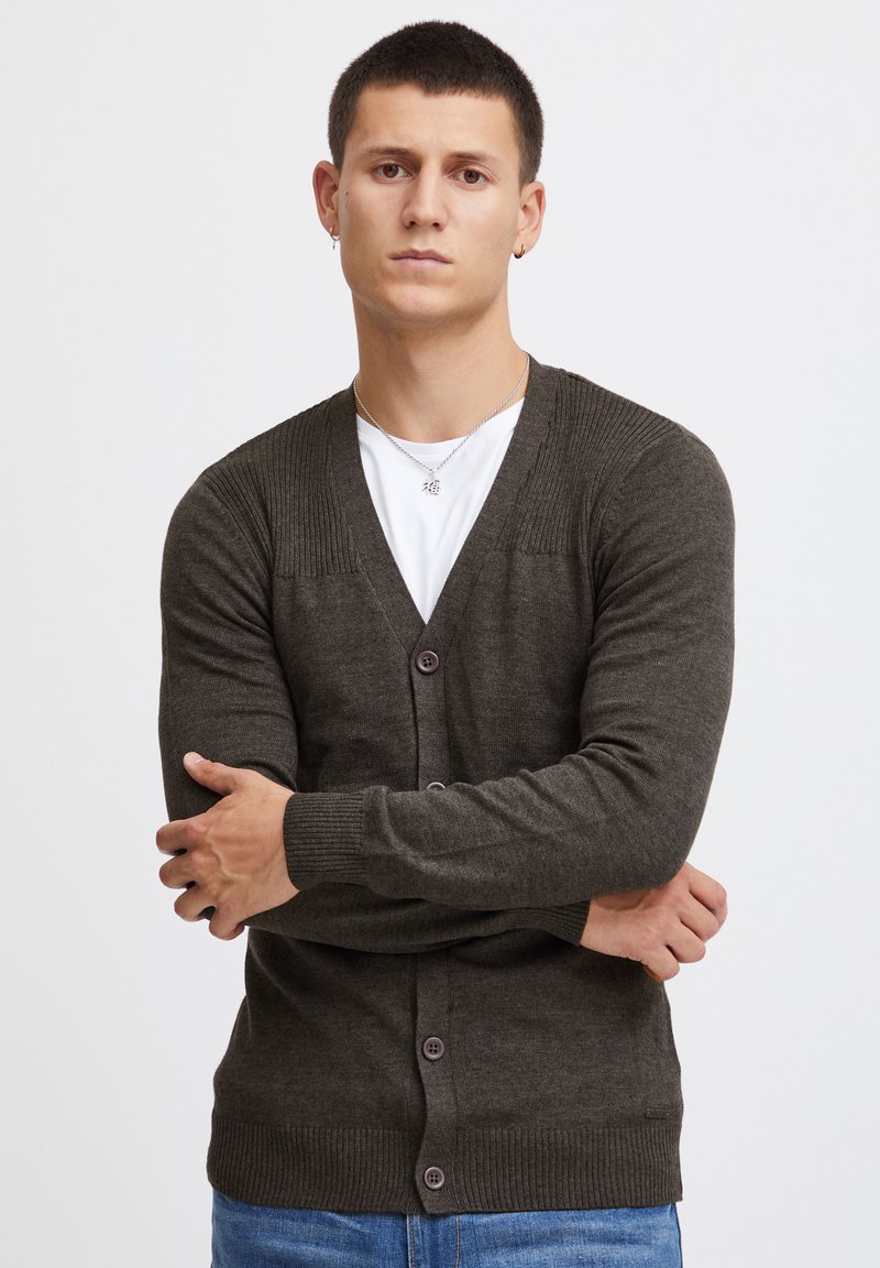 Mørk grønn cardigan med V-hals, ribbet skulderdetaljering, knappelukking og teksturert strikket. Bærer den over en hvit T-skjorte, sammen med blå jeans.