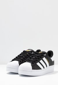 Musta synteettinen urheilukengät valkoisella kärjellä ja pohjalla. Varustettu kolmella valkoisella raidalla, mustilla nauhoilla, teksturoidulla päällisellä ja kultaisella logolla.