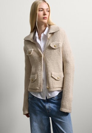 Giacca di maglia beige con colletto a coste, due tasche frontali e bottoni dorati, abbinata a una camicia bianca con colletto e jeans blue.