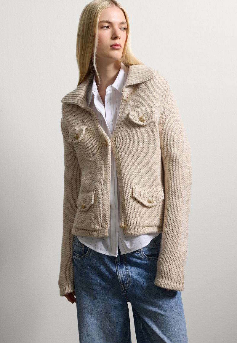 Beige knitted jacket with a ribbed collar, two front pockets, and gold buttons, paired with a white collared shirt and blue jeans.