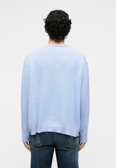 Personne aux cheveux bouclés et foncés, portant un pull bleu clair et un jean bleu foncé, debout les mains relâchées sur les côtés, de dos.