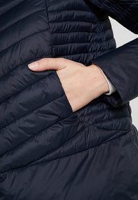 Chaqueta acolchada azul marino con diseño de acolchado horizontal; cuenta con un bolsillo lateral con cierre de cremallera y una textura suave y brillante.