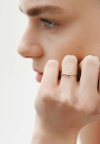 Anillo de oro con una piedra preciosa clara en el centro, sostenido entre los dedos, que muestra una textura suave y un sutil brillo contra un fondo claro.