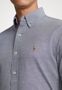 Mężczyzna w jasnoniebieskiej koszuli z guzikami, wykonanej z tkaniny o fakturze, z małym haftowanym logo z polo po lewej stronie klatki piersiowej.