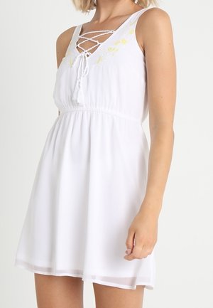 Frau trägt ein ärmelloses weißes Kleid mit gekreuzter Schnürung am Ausschnitt und zarter gelber Blumenstickerei auf der Brust.