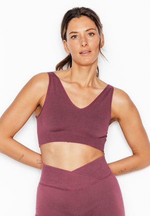 Donna in forma con i capelli legati che indossa un top corto viola senza maniche e leggings a vita alta, in piedi con le mani dietro la schiena.
