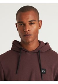 Sudadera color burdeos con una tela texturizada, cordones negros y un pequeño parche con logo en el pecho. Presenta un ajuste casual y relajado.
