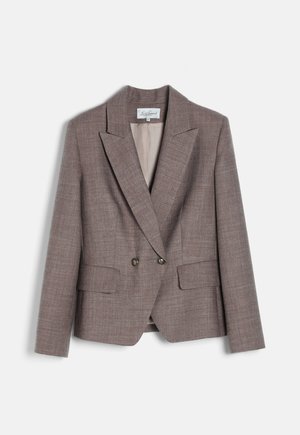 Kvinde iført en brun skræddersyet dragt med bukser med vide ben, hvid bluse, guldbroche, øreringe og armbånd, stående med den ene hånd i lommen og kigger mod venstre.