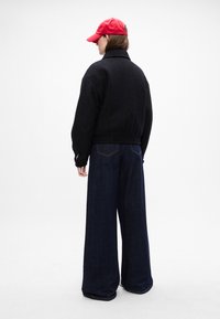 Sort tekstureret jakke med krave, parret med vidde ben mørke denimbukser. Personen bærer en rød kasket og vender væk fra kameraet.