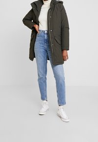 Persoon in een donkergroene winterjack met capuchon, witte coltrui, lichtblauwe jeans en witte hoge sneakers, staand op een effen achtergrond.