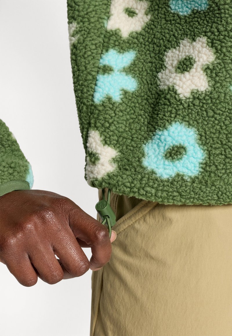 Giacca in fleece verde con motivi floreali in blu e crema. Presenta un orlo regolabile con un toggle verde. Indossata sopra pantaloni in tessuto beige.