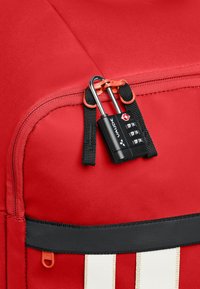 Borsa di tessuto rosso con zip, dotata di un lucchetto combinazione nero e dettagli arancioni. Presenta una tracolla nera e strisce bianche.
