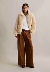 Frau in beigefarbener Sherpa-Jacke, weißem geripptem Top, weit geschnittenen braunen Hosen und braunen Schuhen mit Absatz, vor einem neutralen Hintergrund stehend.