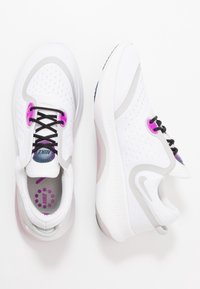 Chaussures de sport blanches avec une tige en mesh, des accents gris, des lacets noirs, et des éléments violets. Semelle semi-transparente avec un motif texturé.