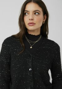 Cardigan noir avec un tricot texturé agrémenté d'accents métalliques, boutons ronds noirs et silhouette ajustée. Porté sur un col roulé noir.